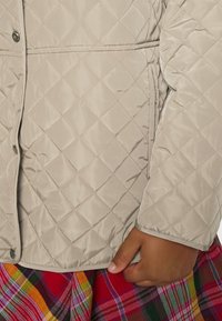 Beige vadderad jacka med en slät textur, med tryckknappar och elastiska ärmslut, matchad med en färgglad rutskjol.