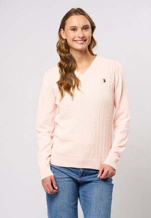 Femme souriante aux cheveux ondulés portant un pull en maille torsadée à col en V rose clair et un jean bleu, debout devant un fond uni.