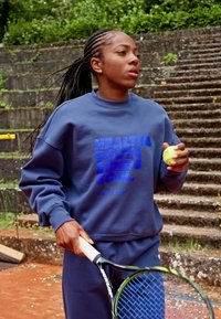 Mörkblå sweatshirt med text, matchande byxor, håller en tennisboll och racket på en lersportplan, med stentrappa och grön bakgrund.