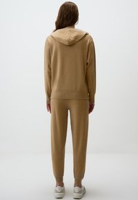 Set di felpa beige e pantaloni jogger realizzato in tessuto morbido, con cappuccio, polsini a coste e vestibilità rilassata. Indossato con sneakers bianche.