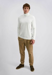 Man in een witte coltrui, bruine corduroy broek en donkerbruine sneakers die op een effen lichte achtergrond staat.