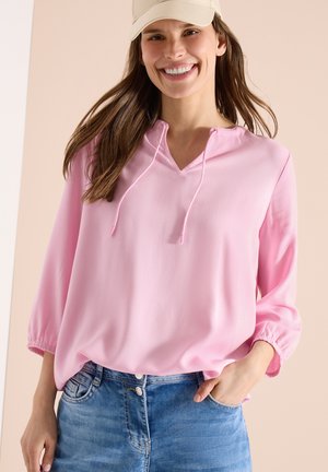 Uśmiechnięta kobieta w beżowej czapce, luźnej różowej bluzce ze sznurkami oraz niebieskich dżinsach, stojąca z jedną ręką w kieszeni.