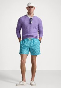 Mężczyzna w purpurowym swetrze w splot kablowy, białej koszuli, niebieskich szortach w wzory, białej czapce, okularach przeciwsłonecznych zawieszonych na swetrze i białych trampkach.