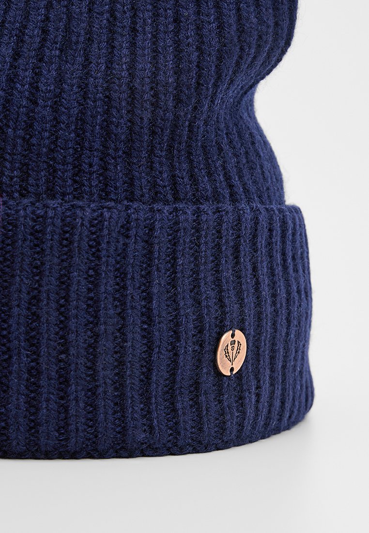 Granatowa dziana czapka z prążkowanym teksturą, z miedzianą metką z detalem logo na zagiętym brzegu.