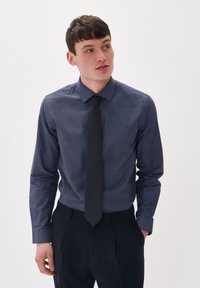 Sötétkék ing apró fehér pöttyös mintával, gombos gallérral, hosszú ujjú, egyszínű sötét nyakkendővel, sötét nadrággal párosítva.