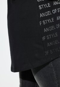 Schwarzes T-Shirt mit leicht abgerundetem Saum, ausgestattet mit einem mattschwarzen Druckmuster mit dem Text "ENGEL DES STILS" an der Seite. Aus Baumwollmaterial.