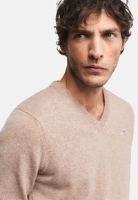 Hellbrauner, V-Ausschnitt-Pullover aus weichem Stoff, mit dezentem Logo auf der Brust und taillierter Form. Strukturierte Oberfläche.