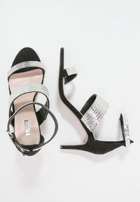 Sandales à talons hauts en suede noir avec des sangles scintillantes argentées, un bout pointu et une fine bride de cheville avec fermoir à boucle.