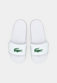 Sandales à enfiler blanches avec une sangle texturée ornée d'un logo crocodile vert. Semelle lisse pour le confort, design minimaliste, sans accents supplémentaires.