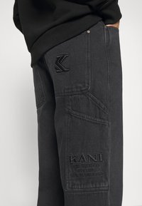 Svarta denim cargo byxor med flera fickor, texturerad tyg och broderad logotyp på benet. Har en distinkt logotypdetalj.