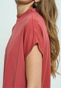 T-shirt à manches courtes couleur rouille, avec un col montant, une texture douce et une coupe décontractée. Détails des coutures aux épaules avec une finition lisse.