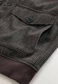 Brown and gray houndstooth patterned fabric with two black buttons and a brown ribbed hem. Features a chest pocket with flap.