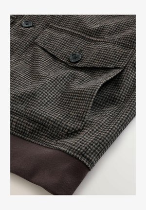 Tessuto marrone e grigio a motivo pied de poule con due bottoni neri e orlo a coste marrone. Dotato di tasca sul petto con pattina.