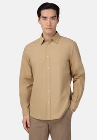 Světle hnědá košile s dlouhým rukávem, s límečkem na knoflíky, předními knoflíky a hladkým, strukturovaným materiálem. V kombinaci s béžovými kalhotami.
