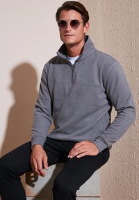 Pullover in pile grigio con colletto a mezza zip, polsini a coste e una cucitura orizzontale in contrasto. Indossato con pantaloni neri.