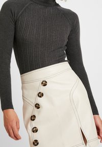 Femme portant un pull gris foncé à col roulé côtelé et une jupe en cuir crème avec de grands boutons en écaille de tortue sur le côté et une fente devant.