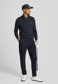 Navy zip-neck pullover, black cap with logo, navy pants, and white shoes with red and blue accents. Model holds a golf club, posing.