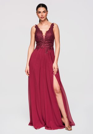 Femme debout portant une robe bordeaux sans manches avec corsage en dentelle et fente haute à la cuisse, associée à des sandales à talons hauts dorées.