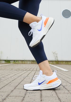 Nike futócipő, főként fehér színben, lila swoosh-zsal és narancssárga részletekkel. Texturált szövet felsőrésze és párnázott talpa van.