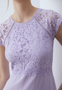 Lilafarget kjole med en blondebisk med blomsterdesign og bølgete kanter, korte ermer og en glatt, flytende skjørt.