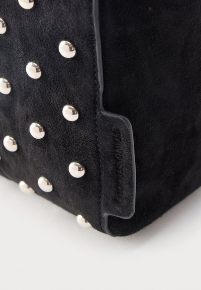 Γωνία μαύρης σουέντ τσάντας χειρός με ασημί στρογγυλά τρουκ και εμφανές ανάγλυφο λογότυπο στη λεπτομέρεια της άκρης, πάνω σε λευκό φόντο.