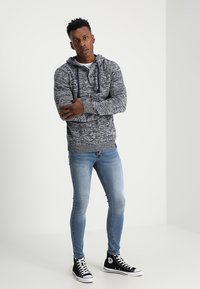 Grijze en witte gemarmerde hoodie met een trekkoord, gecombineerd met aansluitende lichtblauwe jeans en zwarte hoge sneakers. Neutrale achtergrond.