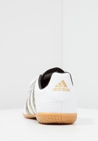 Zapatilla deportiva blanca con parte superior texturizada, acentos negros y tres rayas doradas. Presenta una suela de goma con un agarre con patrón.