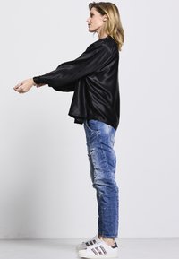 Schwarzes Satin-Oberteil mit langen Ärmeln, lockerer Passform und abgerundetem Saum, kombiniert mit distressed blauen Jeans und weißen Sneakers mit schwarzen Akzenten.