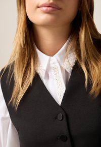 Femeie blondă îmbrăcată cu o cămașă albă cu guler cu mărgele și un veston negru cu nasturi, arătată din gât până la piept.