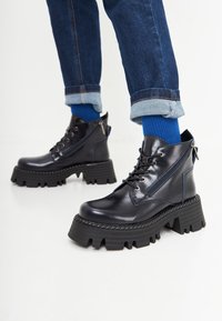 Stivaletti alla caviglia in pelle nera con suola chunky a profondi intagli, cerniera laterale e cuciture contrapposte blu. Indossati con denim arrotolato.