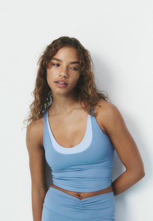 Frau mit lockigem Haar, die ein hellblaues Tanktop und passende Leggings trägt, steht vor weißem Hintergrund und schaut zur Seite.