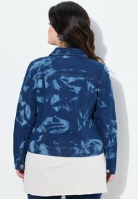 Denimjakke i mørkeblå med tie-dye mønster, med krave, lange ærmer og knapdetaljer ved manchetterne.