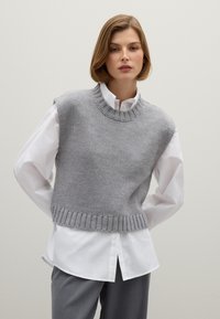 Grauer gestrickter ärmelloser Pullover mit rundem Ausschnitt, mit einem gerippten Saum, der über einem weißen Hemd mit Knöpfen getragen wird. Glatte Textur.