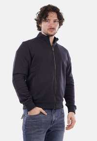 Giacca zip-up blu navy scuro realizzata in tessuto morbido, con tasche laterali, collo alto e polsini a costine, indossata sopra jeans blu.