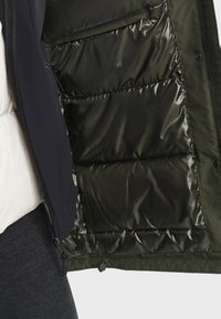 Gestepptes Jacket in Dunkelgrün mit glänzender Innenseite, das einen Reißverschluss und Seitentaschen aufweist. Die Außenseite ist strukturiert und weich.