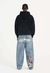 Black hooded sweatshirt with textured pattern; baggy light blue jeans featuring "Ed Hardy" logo and colorful tattoo-inspired graphics.