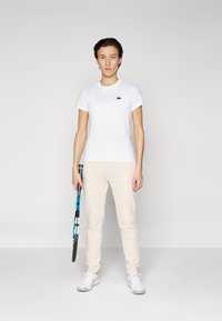 Vit bomullströja med korta ärmar och en mörk logotyp, matchad med ljusrosa avsmalnande sweatpants och vita sportskor, hållande ett tennisracket.