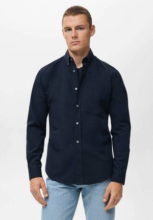 Mørkeblå langærmet skjorte med skjortekrave, forlomme og hvide knapper, parret med lyseblå jeans. Glat stoftekstur.