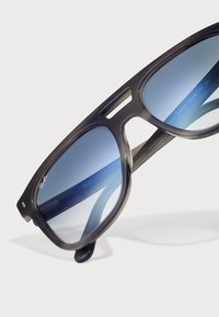 Ochelari de soare Ray-Ban cu un cadru din fâșii de broască țestoasă închise la culoare, lentile cu gradient albastru, design unghiular și un logo proeminent pe lentilă.