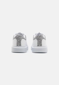 Zapatillas deportivas blancas con un material texturizado, que presentan una atrevida marca negra a lo largo del talón y una suela blanca gruesa. Diseño minimalista.