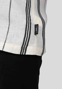 Chemise en lin blanc avec des rayures noires, dotée d'une étiquette sur le côté. Le tissu a une texture douce et une coupe décontractée. Un pantalon noir est porté en dessous.