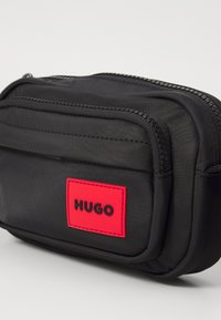 Punguțică din material negru cu două compartimente cu fermoar, având o etichetă dreptunghiulară roșie proeminentă cu "HUGO" scris cu litere negre.