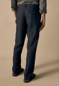 Pantalons bleu marine foncé en tissu texturé, dotés de poches arrière à boutons, associés à des sandales simples.