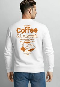 Mann trägt einen weißen Sweatshirt mit orangefarbenem Grafik von einer Kaffeetasse, einem Croissant und Text über Kaffee und Croissants auf dem Rücken.