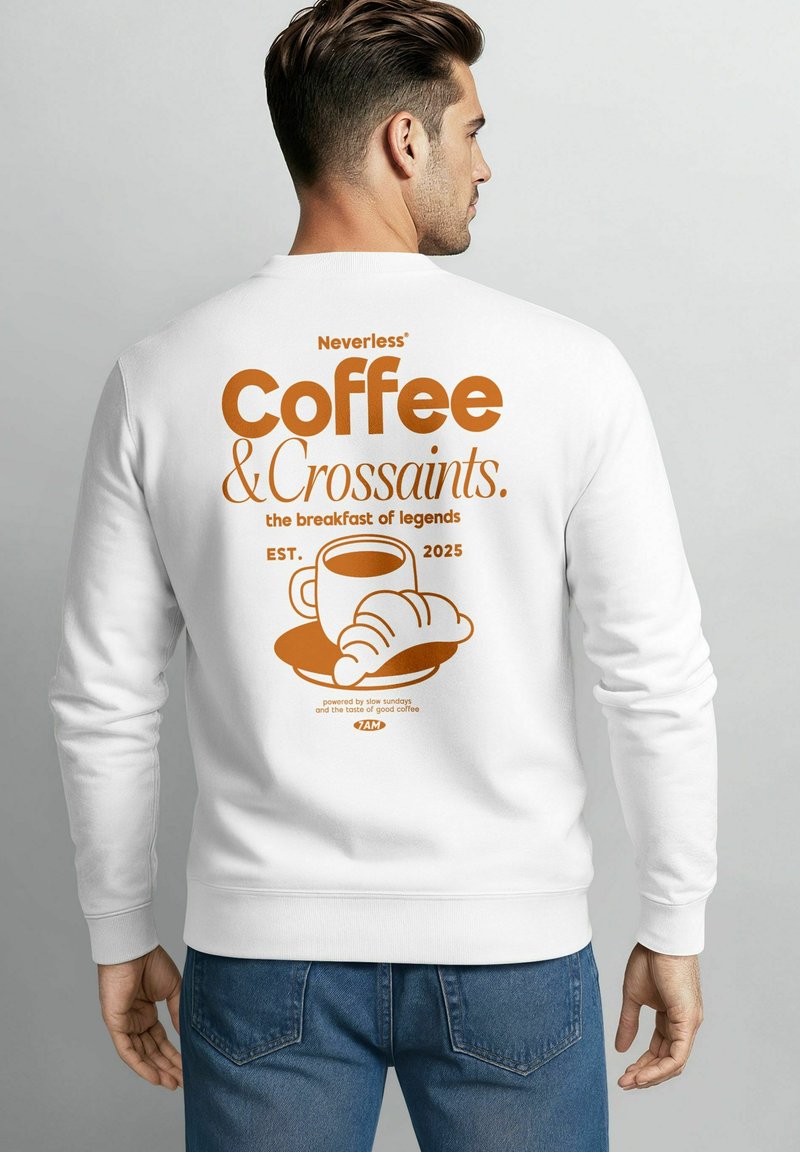 Mann trägt einen weißen Sweatshirt mit orangefarbenem Grafik von einer Kaffeetasse, einem Croissant und Text über Kaffee und Croissants auf dem Rücken.