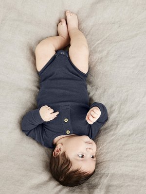 Tumedast sinise soonikuga body pikade varrukatega ja puidust nööpidega, mille seljas on beebi heledat halli kangast taustal. Beebil on lühikesed tumedad juuksed.