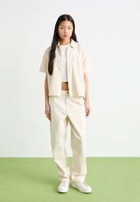 Completo abbinato a righe beige con camicia a maniche corte con bottoni e pantaloni larghi, con una tasca sulla camicia e sneakers bianche.
