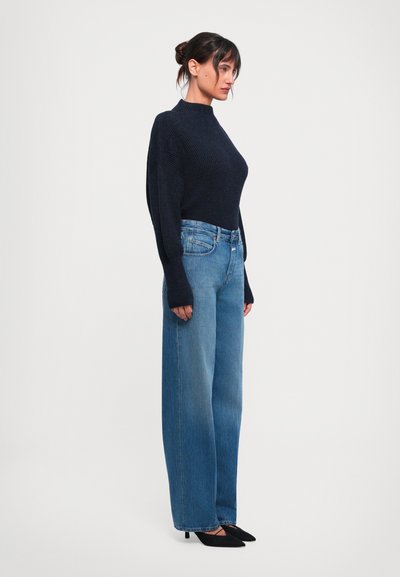 Vrouw in een donkerblauw gebreid sweater, met hoge taille wijde spijkerbroek en zwarte puntige hakken, staand in profiel op een witte achtergrond.