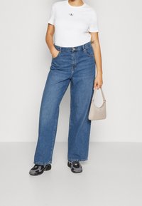 T-shirt bianco leggero con logo, abbinato a jeans blu a vita alta e gamba larga. Gli accessori includono una piccola borsa beige e sneakers nere.