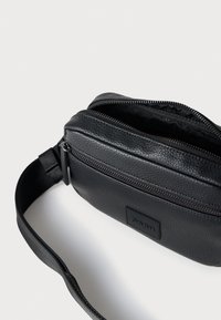 Juodas tekstūruotos odos per petį nešiojamas krepšys su užtrauktuku, reguliuojama dirželiu ir priekyje esančiu etiketės ženklu. Išorinė dalis turi lygią ir grūdėtą apdailą.